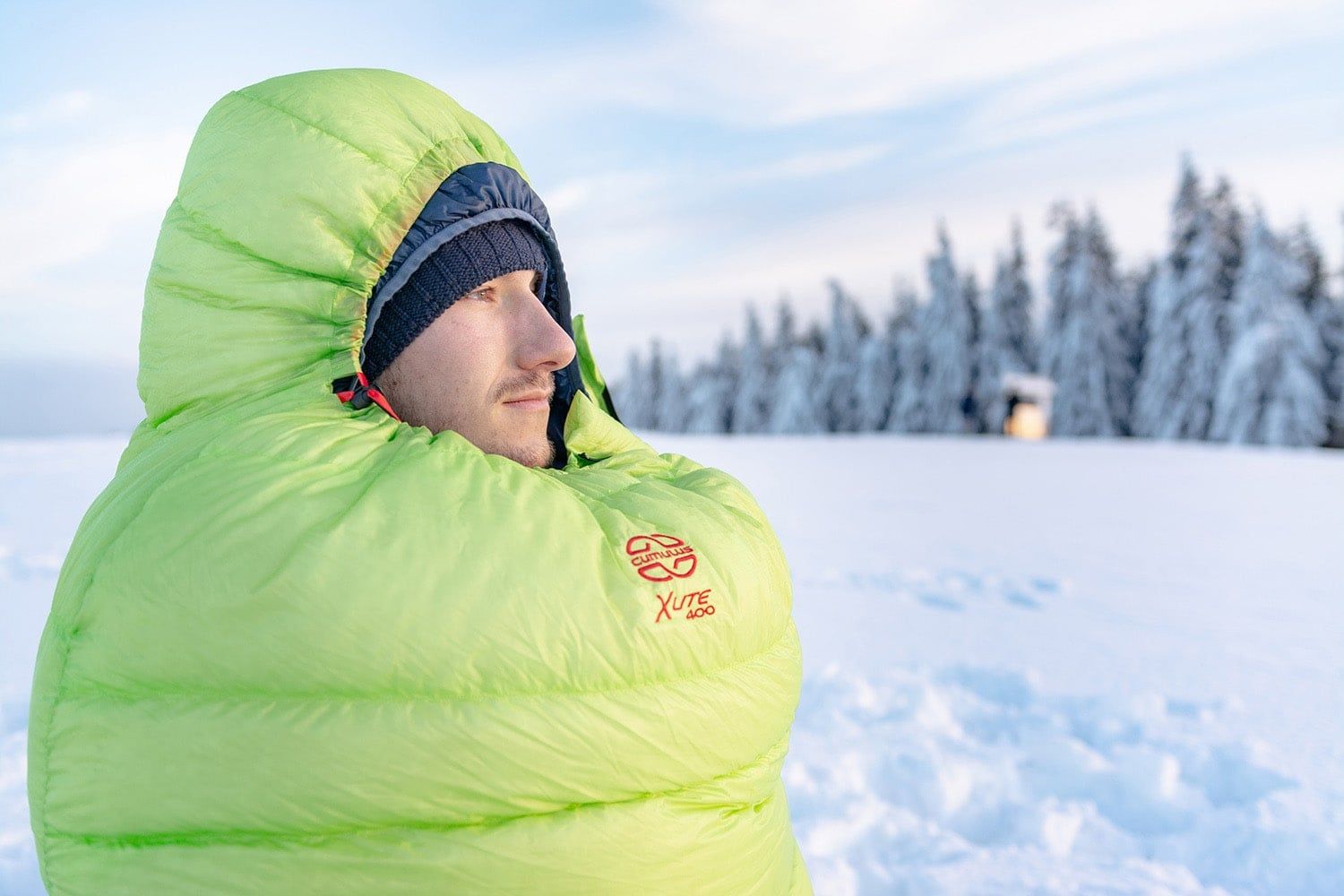 Osoba sedící v péřovém spacáku Cumulus X-Lite 400 se stáhnutým límcem kapuce, v zelené barvě s logem.